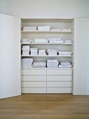 Stacked, white linen on shelves above drawers in white, open cupboard in niche