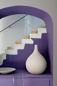 White clay vase artistically arranged on purple cabinet in arched niche in front of staircase