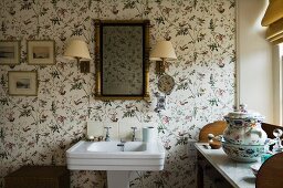 Pedestal sink and mirror on floral wallpaper; porcelain bowl on antique washstand to one side