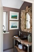Rustikaler Waschtisch unter Badezimmerspiegel mit kunstvollem Rahmen aus Schwemmholz; an der Wand daneben zwei moderne Landschaftsaquarelle