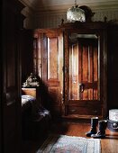 Antiker Kleiderschrank mit Spiegel im Kolonialstil und Stiefel auf Dielenboden in ländlicher Schlafzimmerecke
