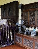 Antique fan and cameras on a carved wooden bench with high back next to a bed with a dark wood headboard and shimmering, violet bed coverlet