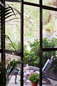Blick durch geöffnete Fenstertür auf Holzterrasse mit Sonnenliege und Eingrenzung durch Grünpflanzen