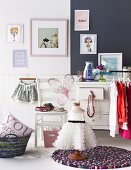 White child's dress on old tailor's dummy standing on round rug in front of chair and chest of drawers below framed pictures on wall painted half dark and half white