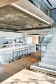 Galley kitchen under exposed concrete ceiling and free standing stairs with glass banister in an open living room