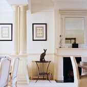 Animal ornament on round side table between open fireplace and classical columns in traditional, white living room
