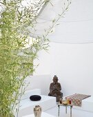 Seating area as continuous, white spatial sculpture half hidden behind bamboo with figure of Buddha and small Oriental tea table
