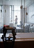 Kettenvorhang hinter Badewanne als Raumteiler zum Schlafzimmer; geschnitzter Holztisch mit Badeutensilien