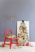 Collagenmix auf weisser Sperrholzplatte als stilisierter Weihnachtsbaum; Geschenkpaket auf rotem Holzstuhl und Stehlampe im Industriestil