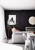 Modern living-dining area in white in front of black wood-panelled wall with white artworks above fireplace and small, antique table