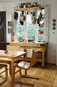 Dining area with Scandinavian-style wooden chairs and wooden table in front of workbench-style shelves below window; pots and pans hanging from ceiling rack