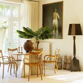 Delicate wooden chairs around modern table and potted banana tree in front of oil painting on wall