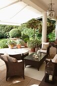 Brown rattan outdoor furniture on a traditional terrace with columns and white awning; view of the garden