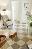 View from a staircase of a dog in an entrance hall with gray and white checkerboard stone floor and upholstered armchair with a floor lamp in a corner
