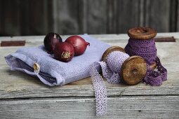 Rote Zwiebeln auf Leinensäckchen neben Holzspulen mit violetter Spitzenbordüre