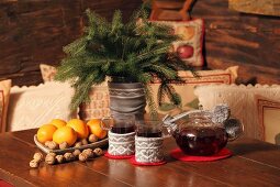 Glasses of tea and fir twigs in vase with knitted cover next to teapot and festive dish of fruit and nuts on wooden table in chalet