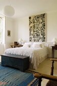 Bedroom with rustic wooden trunk at foot of double bed with lace bedspread and large wall panel with floral motif