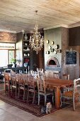 Long dining table and various chairs on Oriental rug below chandelier in rustic dining room with fireplace