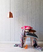 Stack of cushions on wooden chair and pendant lamp with copper lampshade in front of wooden wall