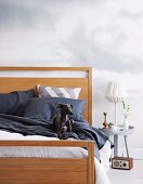 Hund auf dunkler Bettwäsche im Doppelbett mit Holzgestell neben weißem Nachttisch und Tischleuchte