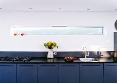 Long, designer kitchen counter with blue base units against wall with narrow ribbon window