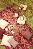 Kissen und Teller mit Speisen auf Picknick-Decke im Ethnolook auf der Wiese