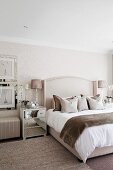 Bedroom in shades of beige with scatter cushions and fluffy blanket on double bed and bedside cabinet with mirrored drawers