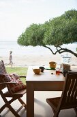 Two wooden chairs and table set for breakfast on terrace with view of tree canopy and ocean beach