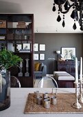 Esstisch mit Silbergerät auf geflochtenem Set; Wohnraum mit Kunstwerken und eleganten Möbeln vor dunkel getönter Wand