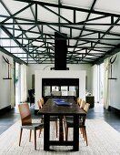 Rustikale Tafel aus dunklem Holz und Stühle im Retro-Stil auf Teppich in hallenartigem Wohnraum mit sichtbarer Dachkonstruktion; Raumteiler vor Loungebereich