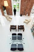 View from gallery down into loft apartment with brick walls, black chairs at large glass table and armchairs arranged casually on purist white floor