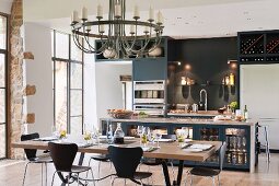 Modern, designer, fitted kitchen with lamps on dark back wall in converted barn with dining table and round chandelier