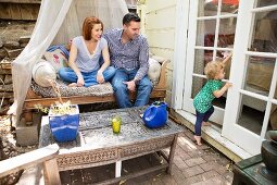 Eltern beobachten kleine Tochter; altes Sofa und geschnitzter Holztisch als ungewöhnliche Terrassenmöbel