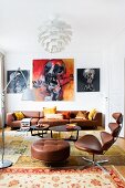 Klassiker Bürosessel mit braunem Lederbezug und passendem Fussschemel vor Loungebereich mit schwarzen Couchtischen und braunem Ledersofa, an Wand expressive Gemälde