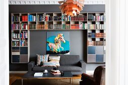 Coffee table and grey sofa in front of modern bookcase and picture on grey wall; Poulsen pedant lamp in foreground