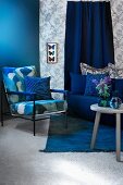 Sitzecke in Blautönen mit Polstersessel, Sofa und Beistelltisch; Schaukasten mit präparierten Schmetterlingen an Wand mit gemusterter Tapete