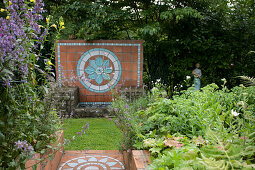 Steps paved with terracotta tiles amongst flowering perennials and back wall of stone fountain with flower-shaped pattern of glazed ceramic tiles