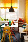 Colourful painted chairs and white bench at dining table with colourful drinking glasses; half-closed, yellow roller blind with scalloped border on window