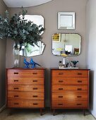 Vase of leaves on one of two wooden chests of drawers against pale grey wall with collection of vintage mirrors