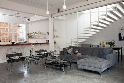 Moderner, offener Wohnraum mit Betonestrich, Wassily Stühlen und großer Couch mit Recamiere; im Hintergrund ein luftiger, breiter Treppenaufgang