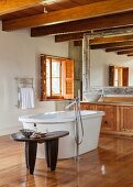 Designer bathtub in spacious bathroom with ethnic table and wooden washstand below wood-beamed ceiling