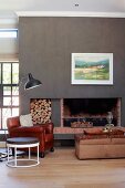 Round side table, leather armchair and retro standard lamp in front of open fireplace with firewood stacked in niche in wall painted dark grey