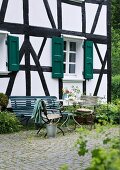 Summery atmosphere on terrace with garden table, garden chairs and bench in front of half-timbered house with green shutters