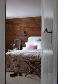View through open door into bedroom with floral bedspread on bed against rustic wooden wall