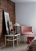 Corner of rustic room with wooden wall, table lamp and framed pictures on side table next to Rococo-style armchair with red and white checked upholstery