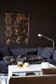 Gemüliches dunkelgraues Polstersofa mit Kuscheldecke vor brauner Wand und Teekanne auf weißem Couchtisch