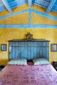 Farb- und Stilkontraste zwischen hellblau gestrichener Holzdecke und ocker marmorierter Wand als Hintergrund für das lasierte Kopfteil eines Doppelbettes