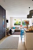 Schwarzweiss gestreifter Läufer vor der Küchentheke und hellblaue Metallhocker im industriellen Vintagestil als Kontrast zu den schlichten Naturtönen des Wohnzimmers