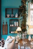 Glaskerzenhalter, Tischlampe und Windlichter auf Tisch, im Hintergrund Holzkästchen mit Weihnachtsdeko und Tannengirlande mit Lichterkette an türkisfarbener Wand