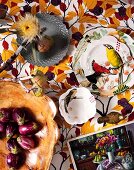 Various dishes made from different materials, some painted with bird motifs and dried flowers on fabric with pattern of autumn leaves